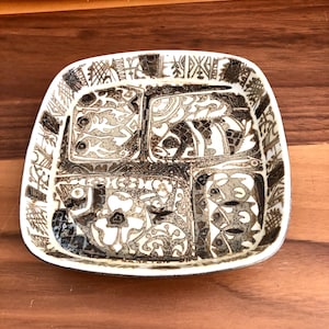 May include: A square, off-white ceramic dish with a brown, fish-themed design. The dish is divided into four sections, each featuring a different fish illustration. The border has a decorative pattern. The dish is on a wooden surface.