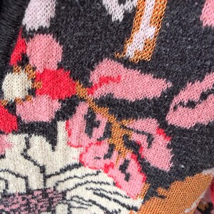 May include: Close-up of a knitted textile with a floral pattern. The design features pink, red, white, and orange flowers on a dark gray background. The knit has a textured appearance, with visible stitches and a soft, cozy look.