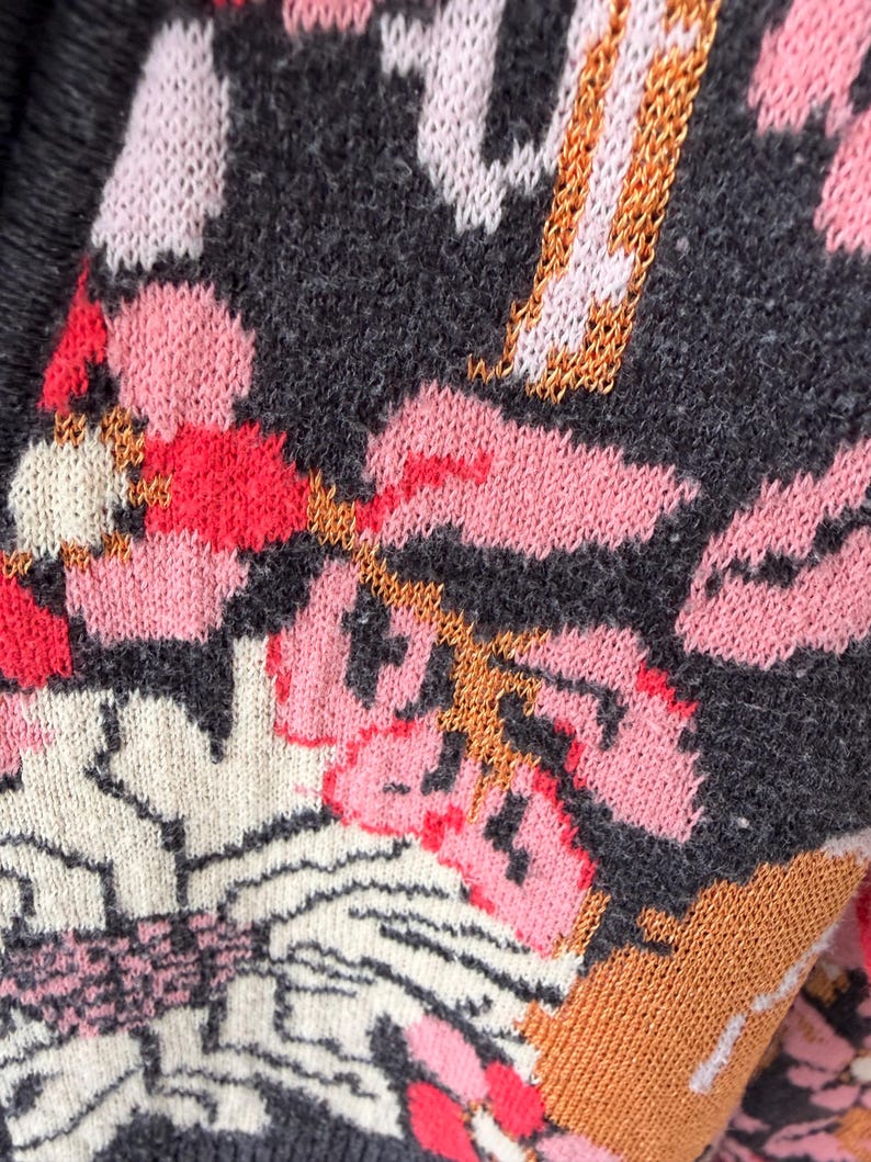 May include: Close-up of a knitted textile with a floral pattern. The design features pink, red, white, and orange flowers on a dark gray background. The knit has a textured appearance, with visible stitches and a soft, cozy look.