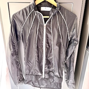 Könnte beinhalten: Eine schwarze Sportjacke mit weißen Paspeln und einem durchgehenden Reißverschluss. Die Jacke hängt an einem Holzbügel. Sie hat einen hohen Kragen und ein kleines Logo auf dem oberen Rücken.