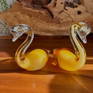 Peut inclure: Deux figurines de cygnes en verre avec des cous incurvés transparents et des corps opaques jaune vif. Les cygnes sont fabriqués avec des surfaces lisses et réfléchissantes et sont placés côte à côte, mettant en valeur leurs cous allongés.