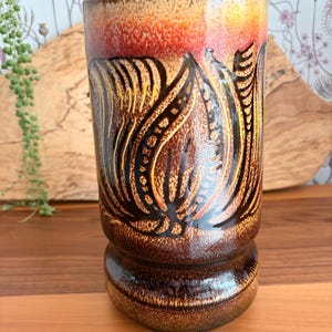 Könnte beinhalten: Eine zylindrische Keramikvase mit einer glänzenden braunen Glasur und strukturierter Oberfläche. Der obere Teil geht in einen leuchtenden Orangeton über. Der Körper ist mit einem dunkelbraunen, eingeritzten Blumenmuster verziert.