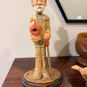 May include: A hand-carved wooden figurine of an elderly man with a white beard and moustache, wearing a tan suit and holding a red object. The figurine stands on a round, wooden base and is holding a walking stick.