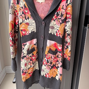 May include: A long, button-down cardigan with a floral pattern in shades of orange, pink, and cream on a black background. The sweater has a gray hood, cuffs, and trim, and two front pockets. The garment is hanging on a wooden hanger.