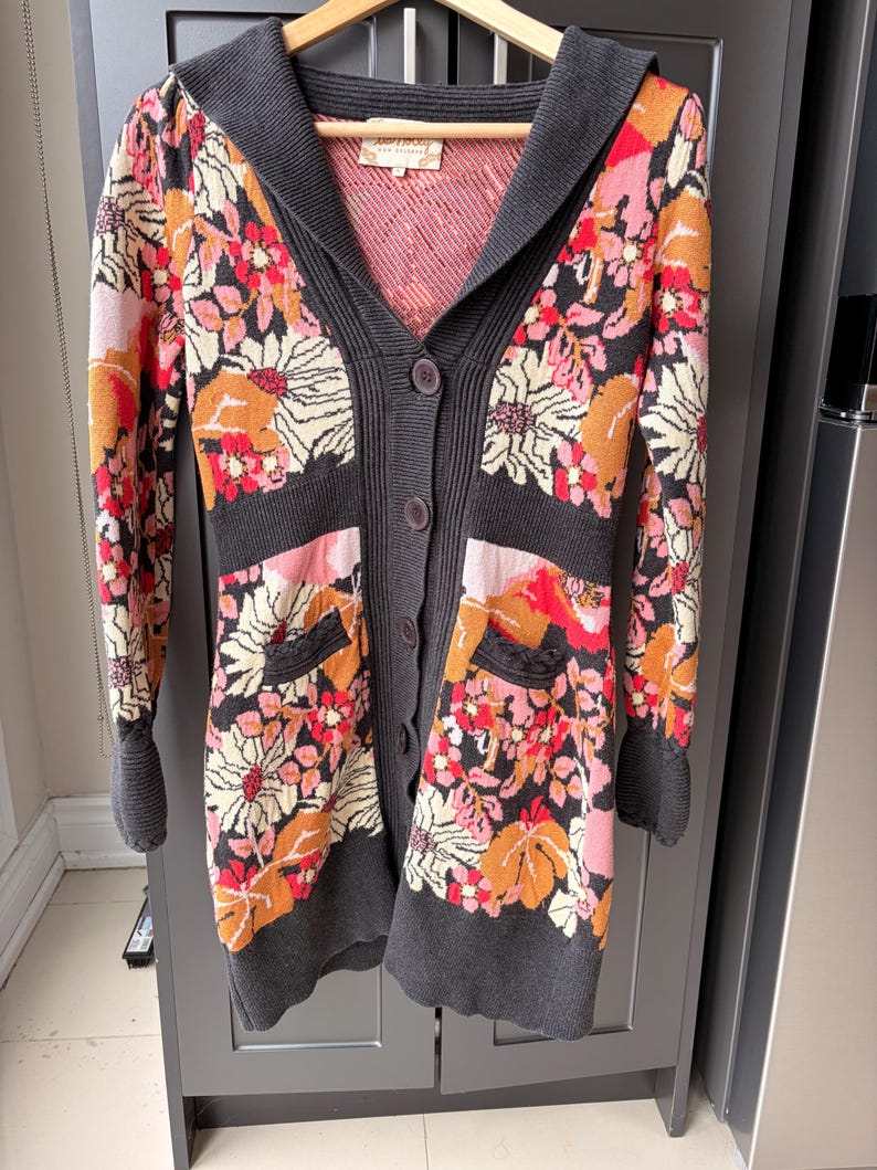 May include: A long, button-down cardigan with a floral pattern in shades of orange, pink, and cream on a black background. The sweater has a gray hood, cuffs, and trim, and two front pockets. The garment is hanging on a wooden hanger.
