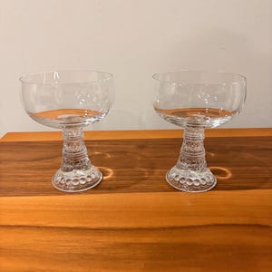 May include: Two clear champagne coupe glasses. Each glass has a wide, shallow bowl and a decorative stem with a series of rings and circular patterns. The glasses are displayed on a wooden surface.