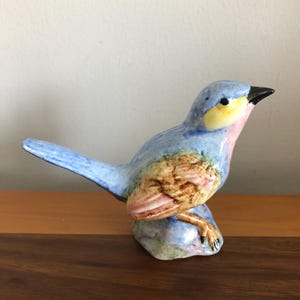 Puede incluir: Figura de pájaro de cerámica en tonos azules, amarillos, rosas y marrones. El pájaro está posado sobre una pequeña base texturizada, con un pico negro y plumas detalladas. La figura se exhibe sobre una superficie de madera.