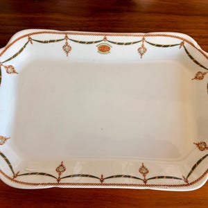 May include: A rectangular white porcelain serving tray featuring a decorative border of green swags, orange beaded chains, and small floral medallions. The centre top displays a small orange oval logo with the text O'NEILL NEW YORK RAILROAD.