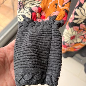 May include: Close-up of a sweater sleeve featuring a floral pattern in shades of pink, orange, and cream. The cuff is a ribbed gray knit with a braided trim. The sleeve is partially visible.
