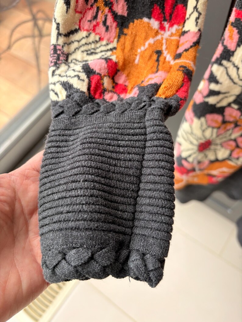 May include: Close-up of a sweater sleeve featuring a floral pattern in shades of pink, orange, and cream. The cuff is a ribbed gray knit with a braided trim. The sleeve is partially visible.