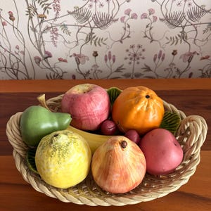 Puede incluir: Una cesta de frutas decorativa de cerámica con un diseño tejido. La cesta contiene una variedad de frutas de cerámica coloridas, incluyendo una manzana, una pera, una naranja y un plátano. El fondo presenta un papel pintado floral.