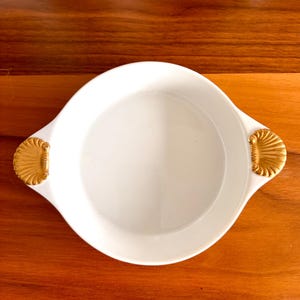 Puede incluir: Un plato de servicio redondo de cerámica blanca con dos asas doradas en forma de conchas marinas. El plato tiene una superficie interior lisa y brillante, con un diseño sencillo y limpio que presenta detalles metálicos en forma de concha.