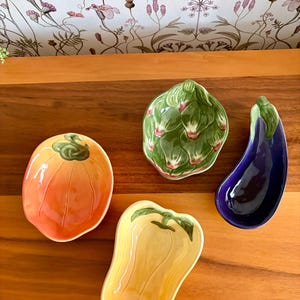 May include: A collection of ceramic vegetable-shaped dishes. The set includes a tomato, artichoke, eggplant, and bell pepper, each with vibrant colors and detailed textures. These decorative pieces are ideal for serving or display.