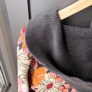 May include: A gray sweater with a floral pattern in shades of pink, orange, and cream. The sweater is hanging on a wooden hanger. The collar is a darker gray.
