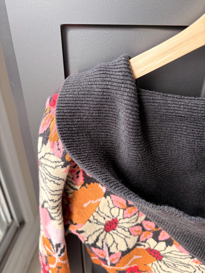 May include: A gray sweater with a floral pattern in shades of pink, orange, and cream. The sweater is hanging on a wooden hanger. The collar is a darker gray.