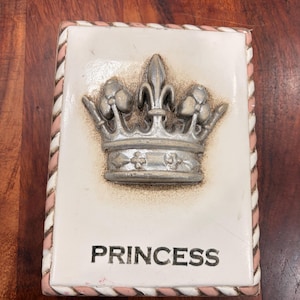 Könnte beinhalten: Rechteckige Keramikfliese mit erhabenem silbernen Kronen-Design und dem Wort "PRINCESS" in Schwarz. Die Fliese hat einen weißen Hintergrund mit einem rosa-weißen Seilrand.