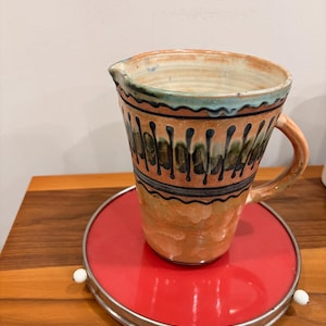 May include: A handmade ceramic pitcher with a handle, featuring a tan base with black and green vertical patterns. The rim has a blue-green glaze. It sits on a round, red metal tray with a silver rim and white accents. The pitcher is on a wooden surface.