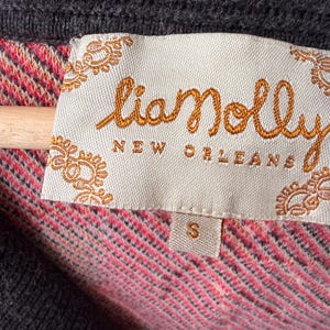 May include: Close-up of a gray sweater with a "lianolly NEW ORLEANS" label. The label is cream-colored with orange embroidered text and decorative edges. The sweater's interior features a pink and red patterned fabric. A small "S" size tag is visible.