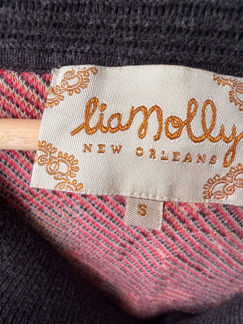May include: Close-up of a gray sweater with a "lianolly NEW ORLEANS" label. The label is cream-colored with orange embroidered text and decorative edges. The sweater's interior features a pink and red patterned fabric. A small "S" size tag is visible.