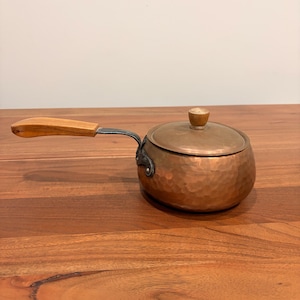 May include: A small copper pot with a lid and a wooden handle. The pot has a hammered texture and a silver-coloured metal attachment for the handle. The lid has a small wooden knob. The pot is on a wooden surface.
