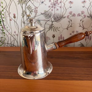 May include: A silver-coloured metal pot with a polished finish, featuring a cylindrical body that flares at the base. It has a small lid with a decorative knob, a short spout, and a long, slender handle made of dark brown wood with ribbed details.