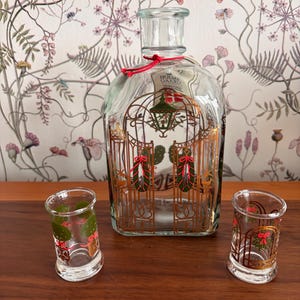 May include: A clear glass decanter and two shot glasses with a gold gate design and red and green accents. The decanter has a red string tied around the neck. The shot glasses and decanter are on a wooden surface.