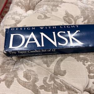 Peut inclure: Une boîte bleu marine de petites bougies coniques DANSK, lot de 12. La boîte porte l'inscription blanche "DESIGN WITH LIGHT". La boîte repose sur un coussin beige à motifs.