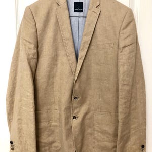 Puede incluir: Una blazer beige con solapa de muesca, cierre de dos botones y dos bolsillos delanteros. La blazer tiene un forro azul claro estampado y está colgada de una percha de madera.
