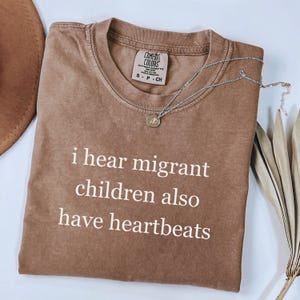 Pu&ograve; includere: Una maglietta Comfort Colors marrone con la scritta bianca "i hear migrant children also have heartbeats". Una collana d'argento con un piccolo ciondolo poggia sulla maglietta. La maglietta &egrave; piegata ed esposta con un cappello e una pianta secca.