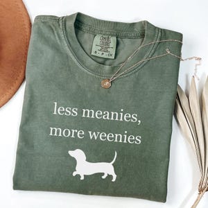 May include: An olive green t-shirt with the text "less meanies, more weenies" and a white dachshund graphic. A gold necklace with a pendant rests on the shirt. The shirt is labeled "Comfort Colors" and is size small.