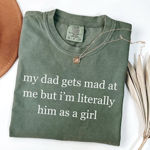 May include: Olive green t-shirt with the text "my dad gets mad at me but I'm literally him as a girl" in white. A gold necklace with a pendant rests on the shirt. The shirt is folded and has a Comfort Colors tag.