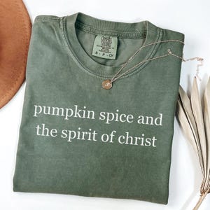 May include: An olive green t-shirt with the phrase "pumpkin spice and the spirit of christ" printed in white. A gold necklace with a pendant rests on the shirt. The shirt is folded and placed on a white surface.
