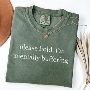 May include: An olive green t-shirt with the text "please hold, i'm mentally buffering" in white. A gold necklace with a pendant rests on the shirt. The shirt is folded on a white surface.