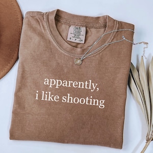 May include: A brown Comfort Colors t-shirt with the text "apparently, i like shooting" in white. A silver necklace with a small pendant rests on the shirt. The shirt is folded and displayed on a white surface.