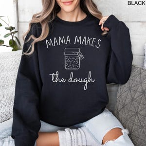 May include: Black sweatshirt with the text "Mama Makes the Dough" in white lettering. A white outline of a jar with a label that says "Dough" is in the center of the text.