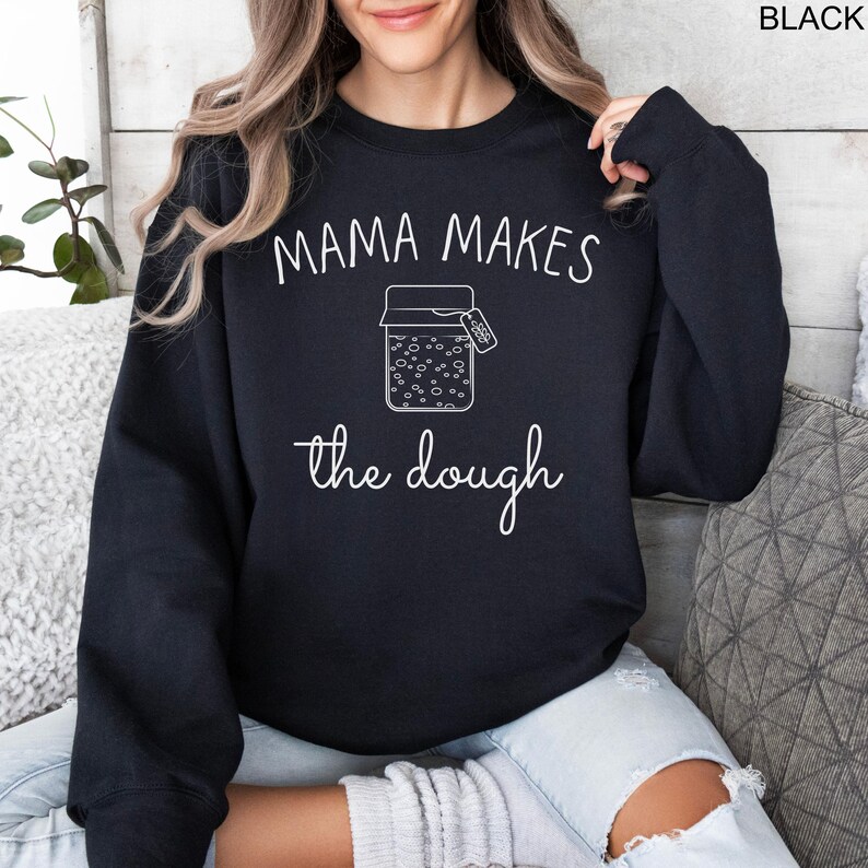 May include: Black sweatshirt with the text "Mama Makes the Dough" in white lettering. A white outline of a jar with a label that says "Dough" is in the center of the text.