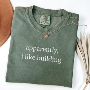 May include: An olive green t-shirt with the text "apparently, i like building" in white. A gold necklace with a circular pendant rests on the shirt. The shirt is a Comfort Colors brand, size S-P-CH.