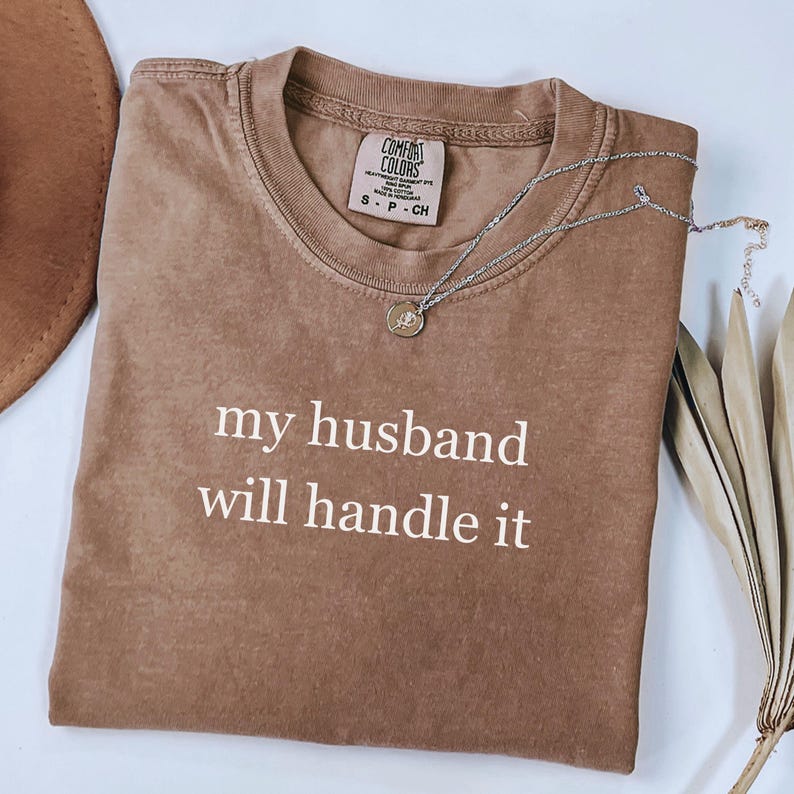 May include: A brown Comfort Colors t-shirt with the text "my husband will handle it" in white. A silver necklace with a pendant is draped across the shirt. A brown hat and dried leaves are in the background.