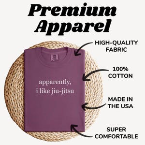 May include: A maroon t-shirt with the text "apparently, i like jiu-jitsu" in white. The shirt is made of 100% cotton and is labeled as high-quality fabric, made in the USA, and super comfortable. The shirt is displayed on a woven surface.