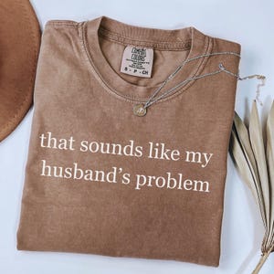 May include: A brown Comfort Colors t-shirt with the text "that sounds like my husband's problem" in white. A silver necklace with a pendant is draped across the shirt. A brown hat and dried leaves are in the background.