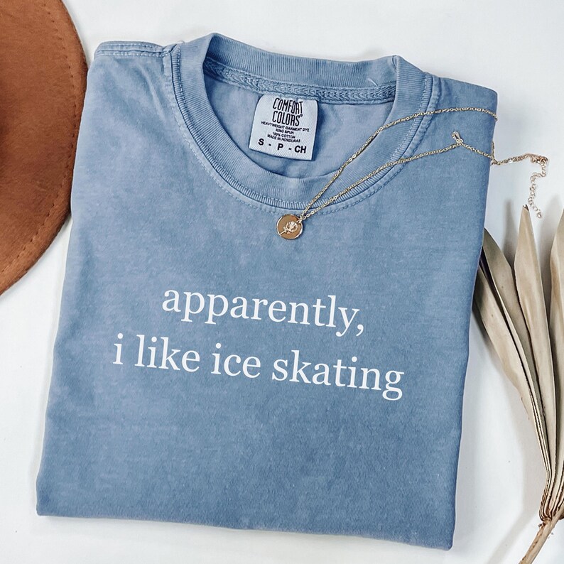 May include: A dusty blue Comfort Colors t-shirt with the text "apparently, i like ice skating" in white. A gold necklace with a pendant and a brown hat are also visible.