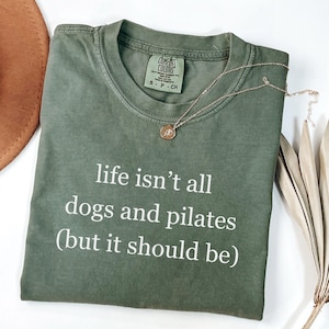May include: An olive green t-shirt with the text "life isn't all dogs and pilates (but it should be)" in white. A gold necklace with a pendant rests on the shirt. The shirt is folded, with a brown hat visible in the background.