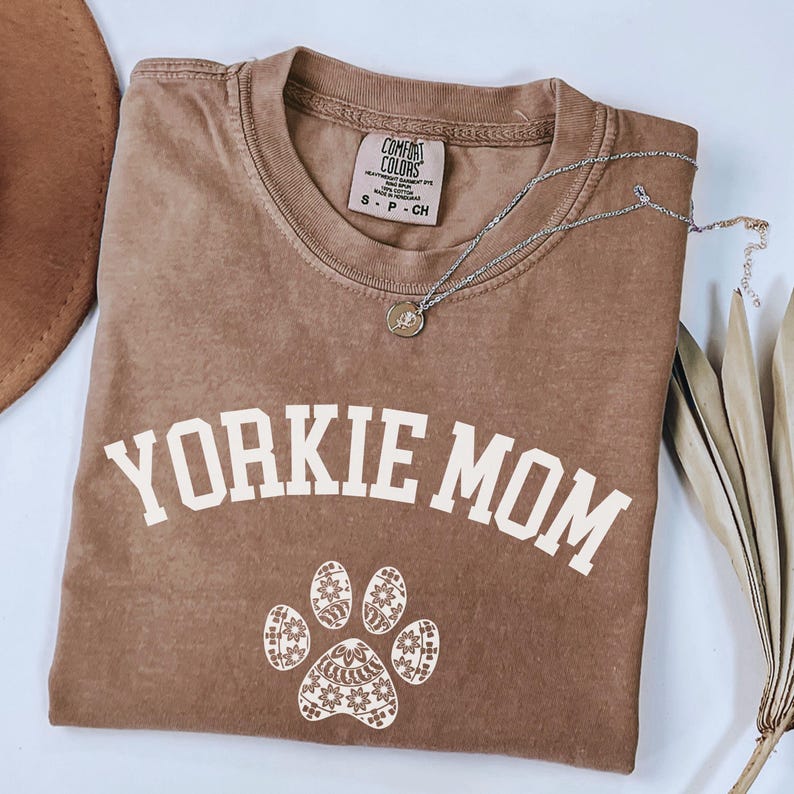 May include: A brown Comfort Colors t-shirt with the text "YORKIE MOM" in white block letters. Below the text is a paw print design. A silver necklace with a pendant is on the shirt.
