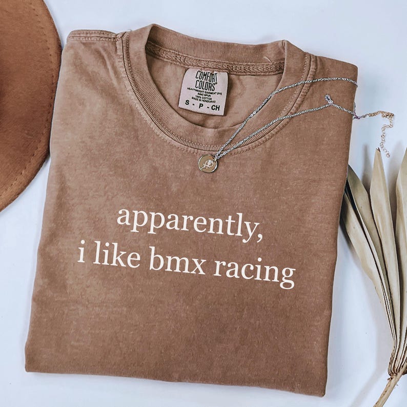 May include: A brown Comfort Colors t-shirt with the text "apparently, i like bmx racing" in white. A silver necklace with a small pendant is draped across the shirt. The shirt is folded on a light surface.