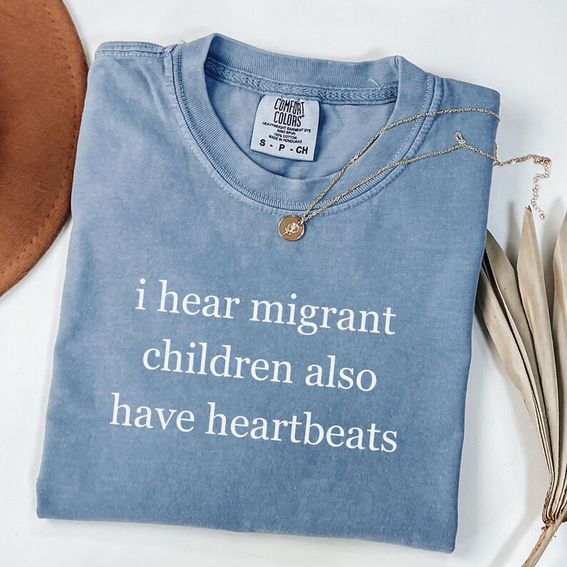 Pu&ograve; includere: Una maglietta blu Comfort Colors con la scritta bianca "i hear migrant children also have heartbeats". Visibile anche una collana d'oro con un ciondolo e un cappello marrone.