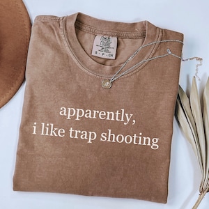 May include: A brown Comfort Colors t-shirt with the text "apparently, i like trap shooting" in white. A silver necklace with a small pendant is draped across the shirt. The shirt is folded on a light surface.