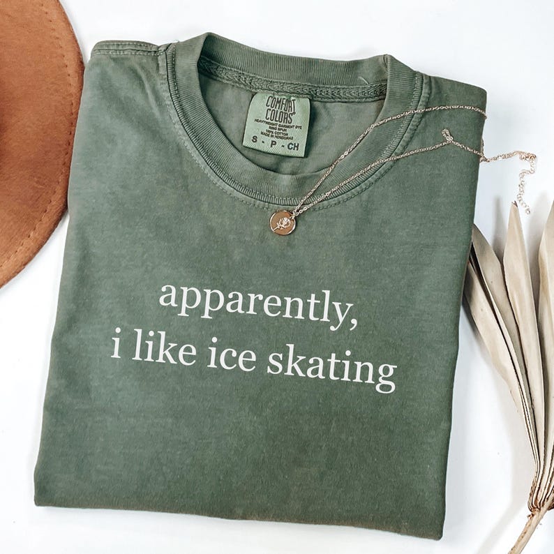 May include: Olive green Comfort Colors t-shirt with the text "apparently, i like ice skating" in white. A gold necklace with a pendant rests on the shirt. The shirt is size S-P-CH.