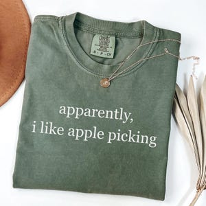 May include: An olive green t-shirt with the text "apparently, i like apple picking" in white. A gold necklace with a pendant rests on the shirt. The shirt has a Comfort Colors tag.