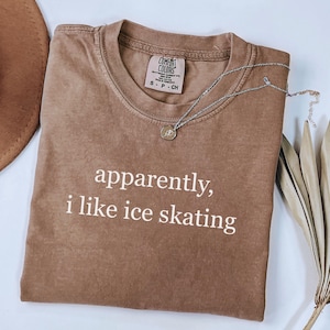 May include: A brown t-shirt with the text "apparently, i like ice skating" in white. The shirt has a crew neck and short sleeves. A silver necklace with a pendant is draped across the shirt.