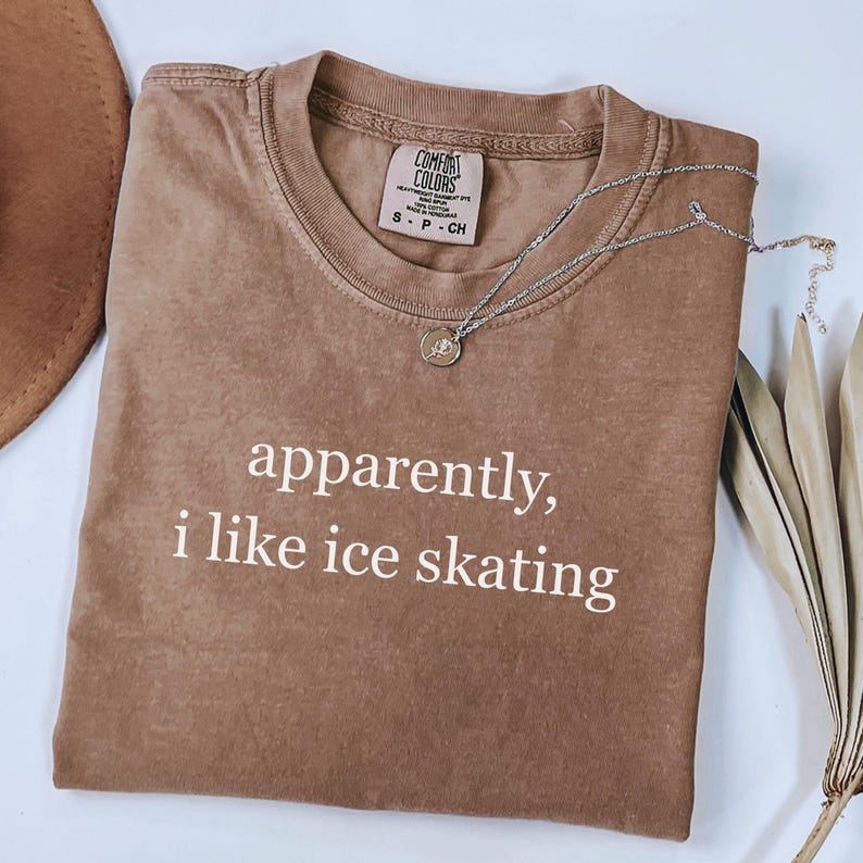 May include: A brown t-shirt with the text "apparently, i like ice skating" in white. The shirt has a crew neck and short sleeves. A silver necklace with a pendant is draped across the shirt.
